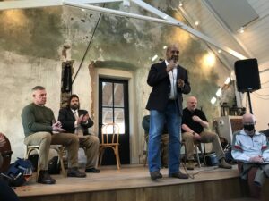 At a Feb 3, 2024 5th District candidates event held at Potter's Craft Cider, candidate Gary Terry, waring a sport jacket and jeans, speaks standing up into into a portable microphone while other candidates and Josh Throneburg listen.