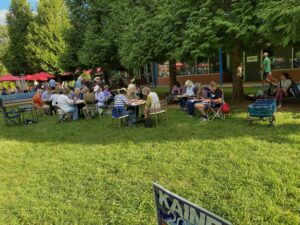 Photo of Cville Dems volunteers working hard in small groups to get out the vote on the grounds of Ix Park on a sunny September day. A Tim Kaine campaign poster is partially visible in the foreground.