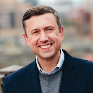 Formal portrait of Ken Martin, 2025 Democratic National Committee chair, wearing a blue button down shirt, gray sweater, and dark suit jacket.