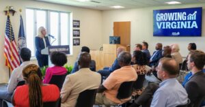 Abigail Spanberger lectures at a meeting titled "Growing Virginia" to a diverse group of seated audience members. American and Virginia State flags are visible at left.