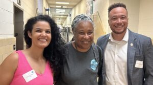Left to right, Delegate Katrina Callsen, Democrat Ivy Hinton, and Delegate Michael Feggans smile in the Carver Rec Center hallway as they enter the Summer Bash. Callsen wears a bright pink dress, Ivy wears a Cville Dems t-shirt, and Feggans wears a plaid cotton jacket over a white shirt.