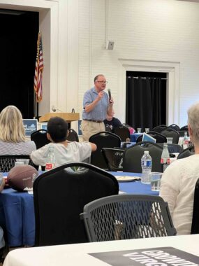 State Senator Creigh Deeds , standing in front of an American flag, rallies the crowd at the Summer Bash 2025. Audience members listen from tables with blue tablecloths.