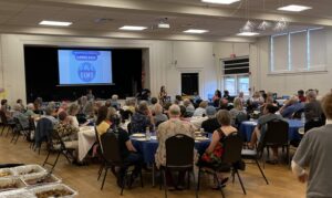 Photo of nearly all attendees at the 2025 Cville Dems Summer Bash. Partygoers are seated at round and rectangular tables with blue tablecloths. A large projection over a stage says "Charlottesville Democrats Summer Bash" and "Cville Dems. Emily Dooley of the Charlottesville school board is speaking to the crowd.