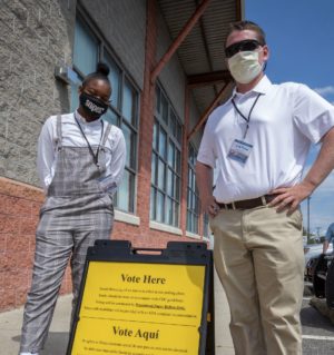 Photo of two masked poll workers â a black woman and a white man â standing next to a yellow sign in English and Spanish