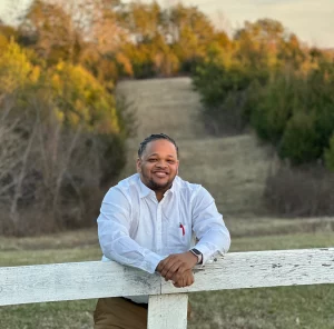 2023 photo of Dashad Cooper in an outdoor setting wearing a long sleeve blue shirt standing behind a white section of fencing.