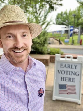 Jeremy Jones with a straw hat and an "I voted" sticker standing in front of an "Early Voting Here" sidewalk sign.