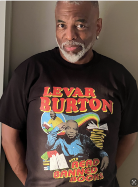 A photo of actor and literacy advocate LeVar Burton wearing a black T-shirt that has two pictures of him, one current and one from his "Reading Rainbow" PBS TV show days (with rainbow graphic) sitting near two stacks of books. The shirt reads "LeVar Burton says read banned books."
