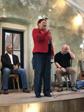 At a Feb 3, 2024 5th District candidates event held at Potter's Craft Cider, candidate Gloria Witt, wearing slacks and a blazer, speaks into a portable microphone while fellow candidate Gary Terry and Josh Throneburg listen.