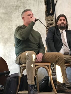 At a Feb 3, 2024 5th District candidates event held at Potter's Craft Cider, candidate Paul Riley, seated and dressed informally, speaks into a portable microphone while fellow candidate Toby Johnson listens.