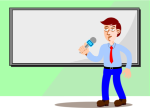 A man wearing a sblue slacks and a blue dress shirt and red tie is talking into a microphone while standing in front of a whiteboard.