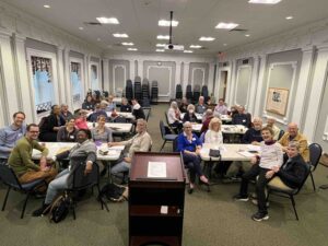 Portrait photo of approx. 30 Cville Dem members seated around 6 rectangular tables in the JMRL Swanson Case room