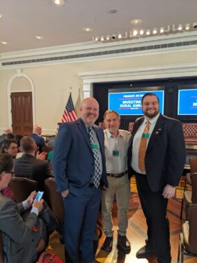 Cville Dems co-chair Josh Throneburg wearing a suit and tie chats with other attendees at the White House rural summit called Investing In Rural America. Television screens and an American flag are behind them.