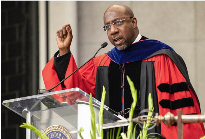 Sen. Reverend Raphael Warnock stands at a podium giving an address wearing a red and black academic robe.