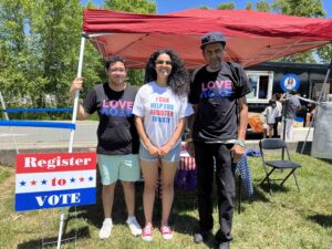 Delegate Katrina Callsen wearing a white "I can help you register to vote" T-shirt is flanked by two male attendees wearing black "Love Vote" T-shirts. All are standing in front of the Voter Registration tent.