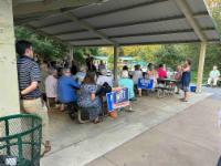 A person addresses the August 2024 meeting of Charlottesville Dems under a picnic shelter. People sit at picnic tables in open air. Witt for Congress and Harris-Walz signs are next to some tables.