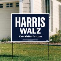 A Harris Walz yard sign with kamalaharris.com at bottom stands in a green lawn in front of a cream-colored house.