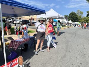 Two smiling women stnad in front of a Cville Dems tent while they work to register voters on a pretty day.