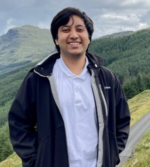 Photo of Kushaan Soodan, wearing a blue collared shirt and a blue jacket, standing in a meadow with hills and forests in the background.