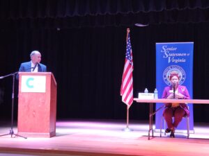 Photo of candidate Gloria Witt seated on center stage behind a table with the moderator looking on from the stage's left side.