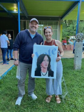 Photo of Josh Throneburg at left and local artist Julia Farill stand together at Ix Park. Julia is holding a poster she created with an original portrait of Kamala Harris; behind Harris’ head is the word "VOTE".