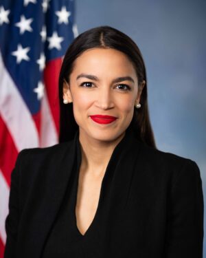 Formal portrait of congressional Rep. Alexandria Ocasio-Cortez, wearing dark suit and standing in front of the American flag.
