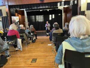 Incumbent James Brown speaks at a stage at Cville Dems Meeting on March 25 2025. Attendees sit on either side of a wooden floor aisle.