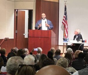 Lamont Bagby gives a speech at redwood lectern a Democratic Party of Virginia meeting to elect an interim leader. He stands next to an American flag in front of a crowd that came to elect the new leader and Bagby won the special election.