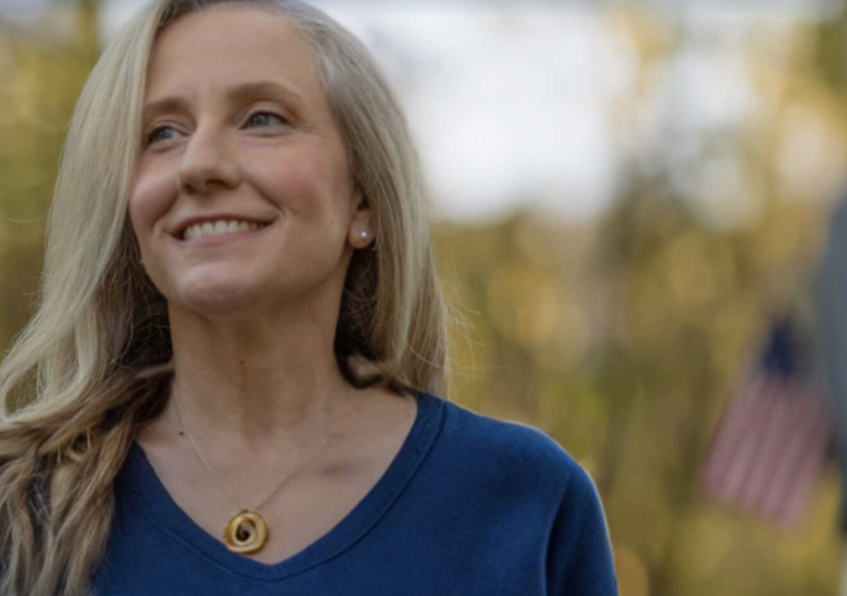 Abigail Spanberger , wearing a blue shirt and gold necklace, smiles widely. In the background is a blurred US flag.